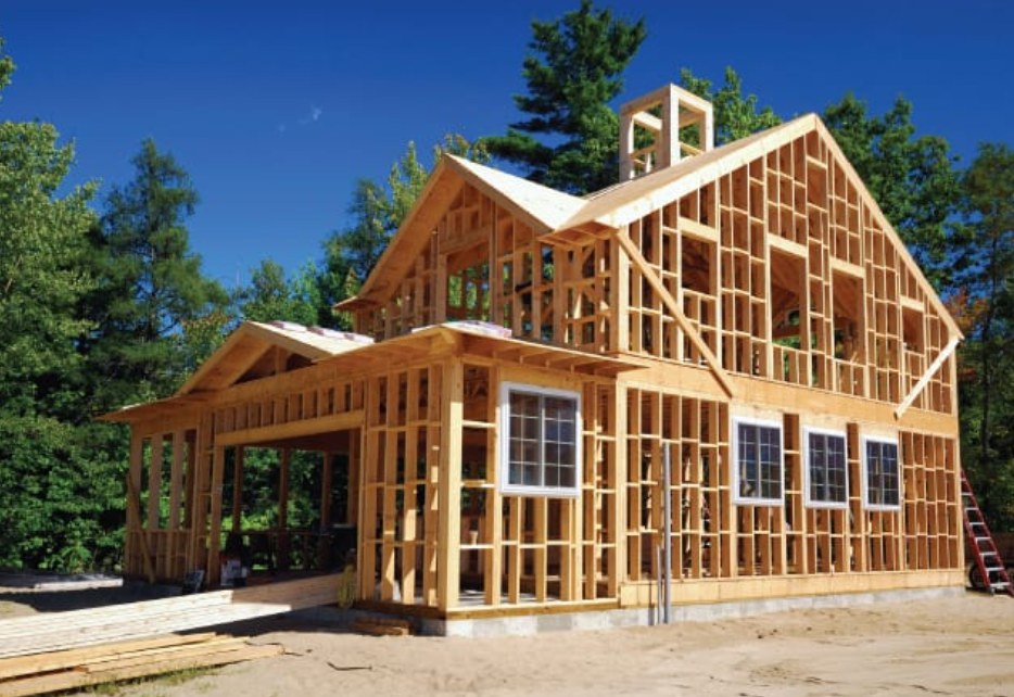 Wood framing of a two-story house under construction beside tall pine trees